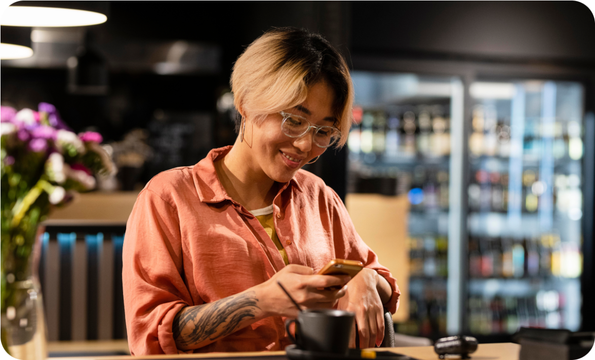 A smiling person with short hair and glasses, wearing a coral shirt, sits at a table in a cozy, modern cafe. They are looking at their phone while holding a coffee cup, with a background of shelves stocked with beverages.