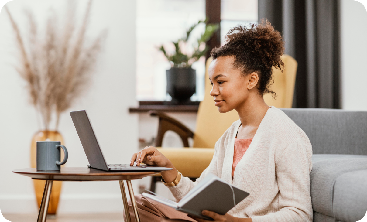 A woman with curly hair, wearing a beige cardigan, sits on the floor in a cozy living room. She is focused on her laptop while holding an open notebook, with a coffee cup placed on a small round table nearby.