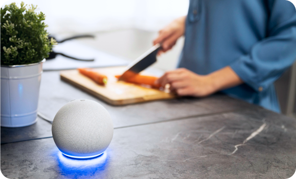 Smart speaker with glowing blue light on a kitchen counter while a person chops vegetables in the background. This image represents Signa Marketing's Voice Search Optimization services, helping businesses adapt to the rise of voice-activated devices by optimizing content and website structure to improve visibility in voice search results.